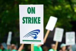 A white sign with green words saying "On Strike" is held up in a strike protest.